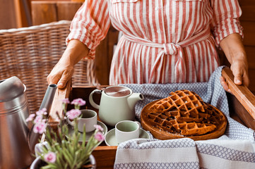 Women holds fresh baked waffles and tea mugs for breakfast. Morning coffee with homemade cakes. Toned photo. Cozy atmosphere at home and flowers on the table.
