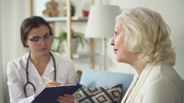 Selective focus on aged woman speaking against doctor in uniform coat. She sitting on comfort couch in bright interior with soft natural light and complains of well-being and health