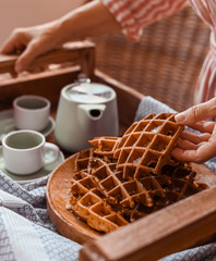 Women holds fresh baked waffles and tea mugs for breakfast. Morning coffee with homemade cakes. Toned photo. Cozy atmosphere at home and flowers on the table.