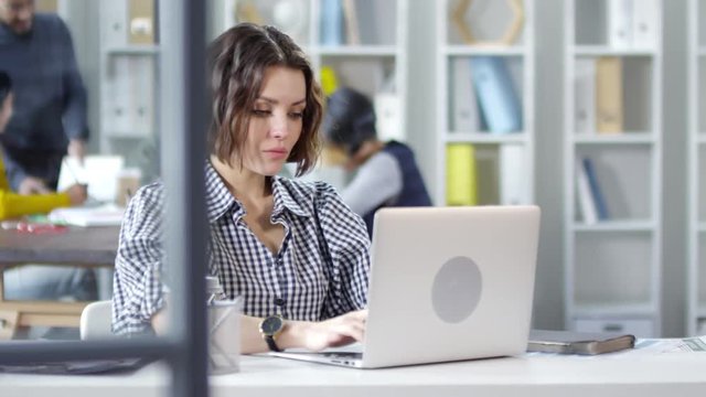 Panning chest-up shot of beautiful young Caucasian girl, wearing plaid blouse, sitting at desk in office, working on laptop then stopping, folding her hands and looking at camera with worried face