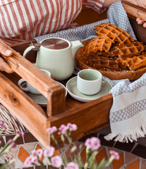 Women holds fresh baked waffles and tea mugs for breakfast. Morning coffee with homemade cakes. Toned photo. Cozy atmosphere at home and flowers on the table.