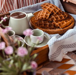 Women holds fresh baked waffles and tea mugs for breakfast. Morning coffee with homemade cakes. Toned photo. Cozy atmosphere at home and flowers on the table.