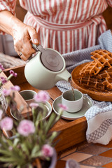 Women holding a kettle and pours tea in a mug. Homemade breakfast with waffles and coffee. Toned photo. Cozy atmosphere at home and flowers on the table.