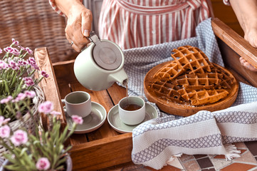 Women holding a kettle and pours tea in a mug. Homemade breakfast with waffles and coffee. Toned photo. Cozy atmosphere at home and flowers on the table.