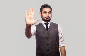 Give me five or stop! Portrait of handsome bearded brunette man in white shirt and waistcoat standing, looking at camera and showing his hand. indoor studio shot isolated on gray background.