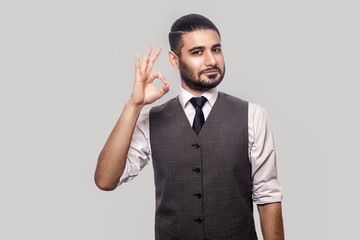 Portrait of happy satisfied handsome bearded brunette man in white shirt and waistcoat standing looking at camera and smiling and showin Ok sign gesture. indoor studio shot isolated on gray background