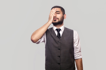 Mistake and sadness. Portrait of sad handsome bearded brunette man in white shirt and waistcoat standing with hopeless face and touching his head. indoor studio shot isolated on gray background.