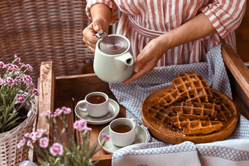 Women holding a kettle and pours tea in a mug. Homemade breakfast with waffles and coffee. Toned photo. Cozy atmosphere at home and flowers on the table.