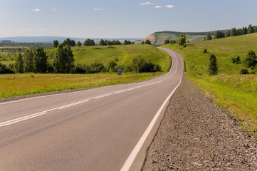 Fototapeta na wymiar Asphalt road going across mountains and green forests. Trees and their shadows on the grass. Sunny summer day with blue sky. Ural landscape