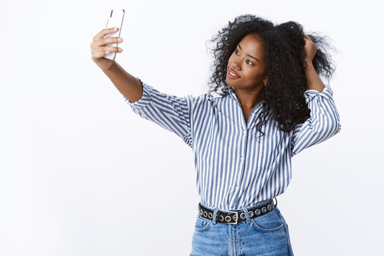 Attractive stylish fashionable confident young charming dark-skinned female blogger playing haircut tilting head flirty showing-off post new pic online holding smartphone taking selfie look display