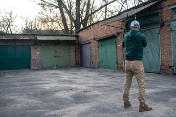 young guy in a white baseball cap shoots a bow. archer is training on the street. a man stands with his back to the camera and shoots a bow.