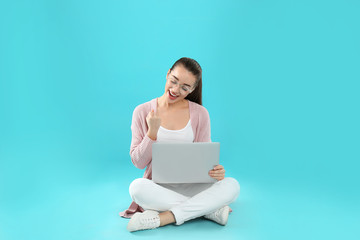 Happy young woman in casual outfit with laptop sitting on color background