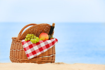Wicker basket with blanket, bottle of wine and fruits near sea, space for text. Beach picnic