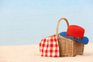 Wicker basket with blanket and hat on sand near sea, space for text. Beach picnic