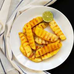 Grilled pineapple wedges on a white plate on a black surface. Summer food. Close-up.