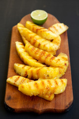 Grilled pineapple wedges on a rustic wooden board on a black background, low angle view. Summer food. Close-up.