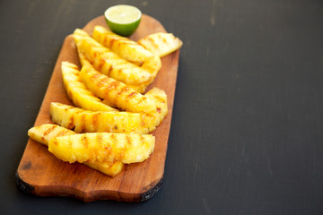 Grilled pineapple wedges on a rustic wooden board on a black background, low angle view. Summer food. Copy space.