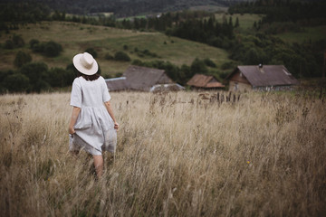 Stylish girl in linen dress and hat walking among herbs and wildflowers in field. Boho woman enjoying day in countryside, simple slow life style. Space for text. Atmospheric image
