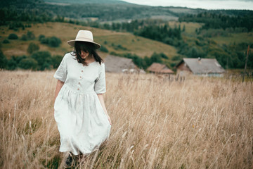 Stylish girl in linen dress and hat walking among herbs and wildflowers in field. Boho woman...