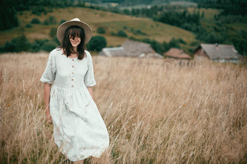 Stylish girl in linen dress and hat walking among herbs and wildflowers in field. Boho woman smiling and relaxing in countryside, simple slow life style. Space for text. Atmospheric image