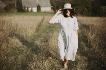 Obraz premium Stylish girl in linen dress and hat walking barefoot among herbs and wildflowers in sunny field in mountains. Boho woman smiling in countryside, simple rustic life. Atmospheric image