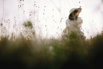 Stylish girl in rustic dress and hat sitting among wildflowers and herbs in sunny meadow  in...