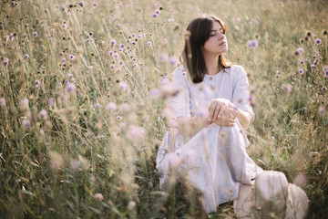 Stylish girl in rustic dress sitting among wildflowers and herbs in sunny meadow in mountains. Boho...