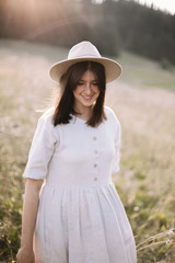 Stylish girl in rustic dress smiling and walking in wildflowers in sunny meadow in mountains. Portrait of happy boho woman in countryside  at sunset, rural simple life. Atmospheric image