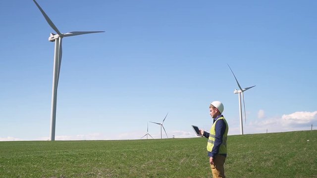 Adult or mature specialist man in protective uniform wear standing with portable tablet in hands against landscape wind turbine power station on background. Male checking information on device