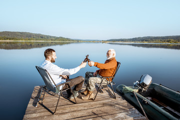 Grandfather with adult son enjoying beer, sitting together on the pier while fishing on the lake early in the morning. Wide landscape view