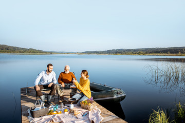 Man and woman with senior grandfather having a picnic with vegetables and fresh caught fish on the lake in the morning. Wide landscape view