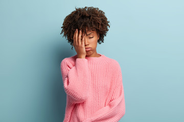 Beautiful overworked woman feels sleepy, covers half face with palm, keeps eyes closed, sighs from tiredness, wears pink sweater, isolated on blue background. Negative feelings and boredom concept
