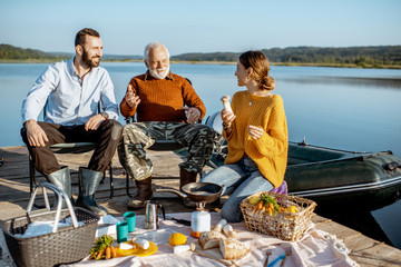 Man and woman with senior grandfather having a picnic with vegetables and fresh caught fish on the lake in the morning