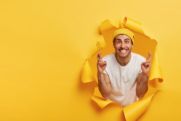 Funny pleasant looking young man waits for special moment, keeps fingers crossed, wishes dreams come true, poses in paper hole, copy space on yellow background. People and body language concept