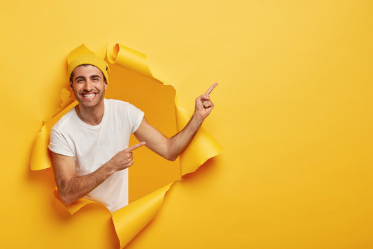 Excited pleased young man in casual outfit, shows something great on blank space, advertises product, poses in torn hole in yellow paper wall, empty area for your advertising content or promotion