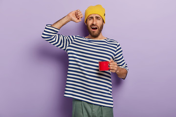 Morning starts with coffee. Sleepy man stretches after waking up, drinks hot beverage to feel refreshed, holds red cup, says good morning, wears casual clothes. People, awakening, drinking concept