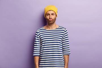 Shot of handsome youngster with bristle pouts lips and looks mysteriously, being dissatisfied with something, wears yellow hat and striped sailor jumper, isolated over purple background, makes grimace