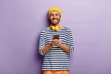 Waist up shof of glad attractive hipster wears stereo headphones and holds modern cell phone, listens audio track and checks notification, dressed in striped sailor jumper, yellow headgear, smiles - obrazy, fototapety, plakaty