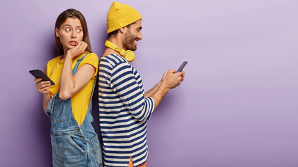 Young couple stand back to back with mobile phones, jealous nervous woman tries to spy what boyfriend is typing, pose against purple background with empty space. Relationship, secrecy, infidelity