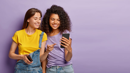 Pleased female friends watch something interesting on mobile phone, have happy relaxed expressions, focused in device, enjoy using modern technologies, isolated over purple wall with empty space