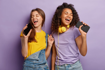 Optimistic pleased mixed race women sing favourite song in smart phones, have fun and enjoy music,...