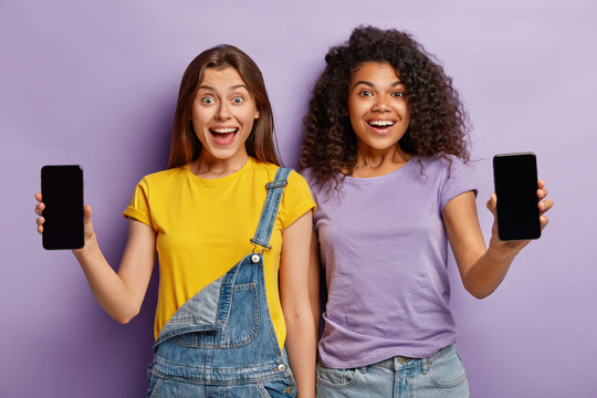 Friendship, technology, advertising concept. Two smiling multiethnic teenagers stand closely, show smartphones with mockup screens for your text, being in high spirit, pose against purple background.
