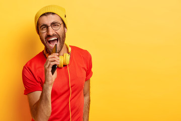 Energetic positive guy sings loudly, keeps mobile phone as if microphone, headphones connected to...