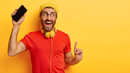 Delighted happy man has fun, dances to music, holds modern cell phone, looks aside, smiles...