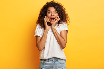 Sociable friendly impressed curly African American teenage girl holds breath from awesome news, calls friend via mobile phone, keeps mouth widely opened, dressed in casual white t shirt and jeans.