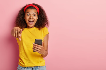 Emotional beautiful Afro female points at camera with wonder, expresses happiness, holds smartphone device, enjoys virtual communication, checks mail, uses free wifi wears yellow casual t shirt