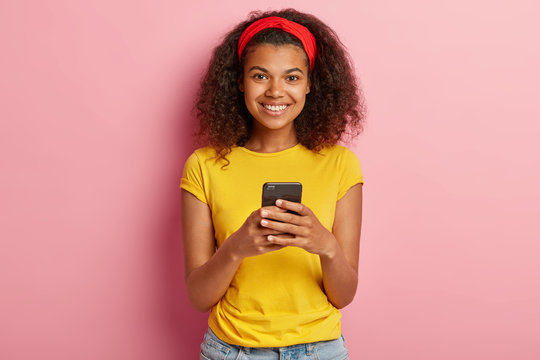 Smiling happy dark skinned teenager reads content text, chats online, wears yellow t shirt, looks happily at camera, blogs in social networks, uses free intrnet connection. Technology and lifestyle