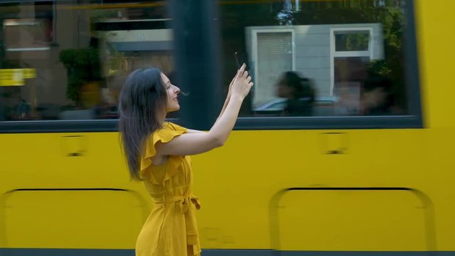 a young charming brunette in a stylish yellow dress takes pictures on the phone in the middle of the street while a yellow tram passes behind her back