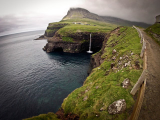 Wide extra panoramic view on Faroe Island famous waterfall with small town above it. Cloudy sky and green hills in the background.