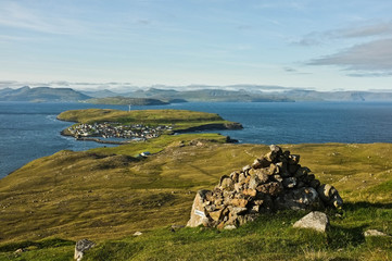 View on multiple islands with small city down the hill. Green grass, blue ocean and cloudy sky in the background. Stone pile placed for orientation purpose in front. Faroe Islands. - obrazy, fototapety, plakaty